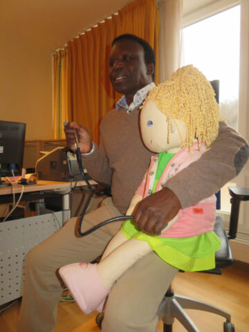 Dr. Paul Okot-Opiro hat Handpuppe Ilona auf dem Schoß und Stethoskop in der Hand